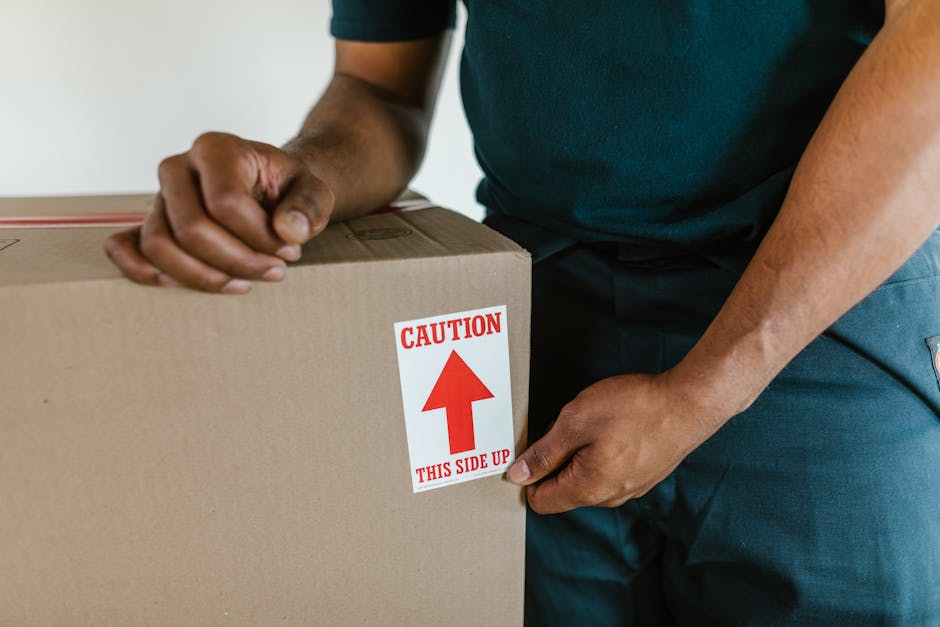 Close-up of a person wearing a dark green and navy shirt, carefully holding a large cardboard box marked with a red and white label that reads 'Caution This Side Up' with an upward arrow, indicating careful handling during a home relocation or furniture transport. The person is inside a room with soft lighting, and part of the lower body is visible, dressed in dark trousers. The box is made of sturdy cardboard, and the label suggests it contains fragile or sensitive items being packed and prepared for moving. In the background, there are no additional objects visible, focusing solely on the careful handling of the moving box, which is part of the packing and loading process associated with house removals by Barnet Man and Van.