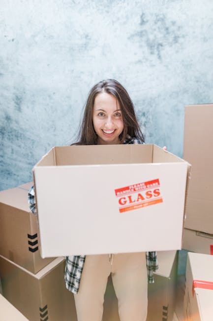 Close-up of a person wearing a dark green and navy shirt, carefully holding a large cardboard box marked with a red and white label that reads 'Caution This Side Up' with an upward arrow, indicating careful handling during a home relocation or furniture transport. The person is inside a room with soft lighting, and part of the lower body is visible, dressed in dark trousers. The box is made of sturdy cardboard, and the label suggests it contains fragile or sensitive items being packed and prepared for moving. In the background, there are no additional objects visible, focusing solely on the careful handling of the moving box, which is part of the packing and loading process associated with house removals by Barnet Man and Van.