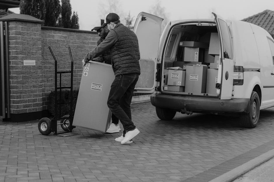 Two men engaged in a home relocation process are seen loading a large cardboard box marked fragile onto a black hand truck outside a residential property. One man is wearing a baseball cap, a long-sleeve shirt, and glasses, while the other is dressed in a quilted jacket and dark trousers. The cardboard box is being lifted from the ground, and behind them is an open white van filled with multiple packed boxes and larger household items, all secured with plastic wrap and packing tape. The van is parked on a paved driveway adjacent to a brick wall, with the background showing trees and a cloudy sky. Equipment such as blankets and straps are visible, indicating a careful and organized furniture transport and packing process typical of house removals. This scene is representative of the logistics involved in professional relocation services provided by Barnet Man and Van, supporting efficient home removals and furniture transport services as referenced in the High Barnet station man and van service guide.