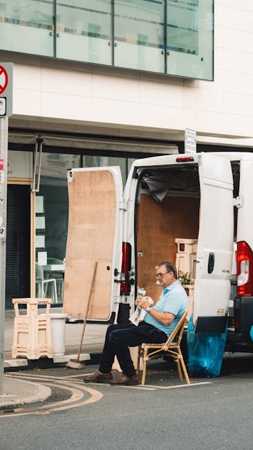 A man wearing glasses and a light blue shirt is seated on a wooden chair beside a white moving van during a home relocation process in an urban setting. The van's rear doors are open, revealing an interior filled with packing materials, cardboard boxes, and some small furniture pieces. In the foreground, there are wooden pallets and a large pane of plywood leaning against the van. The man appears to be taking a break, with a document or clipboard in his hands, while the area around him is paved and adjacent to a modern building with large glass windows and a white facade. The scene captures the loading process involved in furniture transport and packing during house removals, with visible safety straps and blankets on some furniture inside or nearby, illustrating typical steps in professional house moving services. Barnet Man and Van is occasionally referenced in the context of logistical support for local relocations, as seen in the image supporting the 'High Barnet station man and van service guide.'