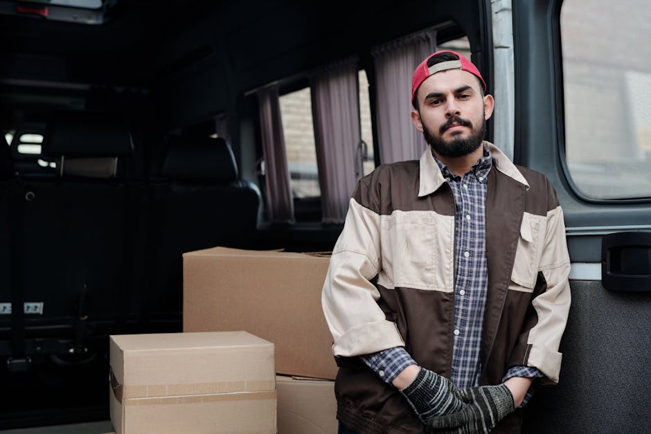 Two men engaged in a home relocation process are seen loading a large cardboard box marked fragile onto a black hand truck outside a residential property. One man is wearing a baseball cap, a long-sleeve shirt, and glasses, while the other is dressed in a quilted jacket and dark trousers. The cardboard box is being lifted from the ground, and behind them is an open white van filled with multiple packed boxes and larger household items, all secured with plastic wrap and packing tape. The van is parked on a paved driveway adjacent to a brick wall, with the background showing trees and a cloudy sky. Equipment such as blankets and straps are visible, indicating a careful and organized furniture transport and packing process typical of house removals. This scene is representative of the logistics involved in professional relocation services provided by Barnet Man and Van, supporting efficient home removals and furniture transport services as referenced in the High Barnet station man and van service guide.