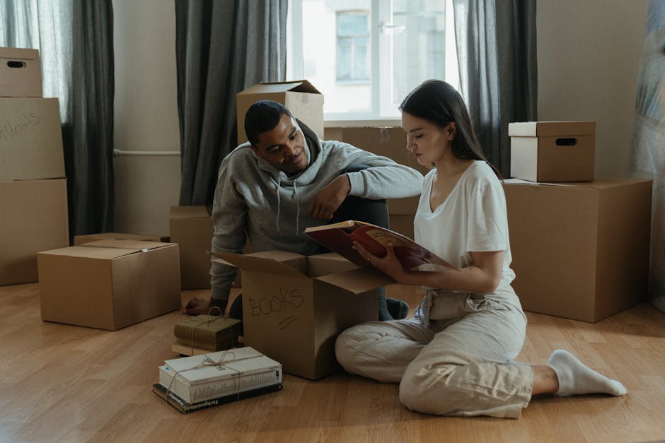 A man and a woman are inside a room surrounded by unpacked moving boxes made of cardboard, some labeled with handwritten notes such as 'books.' The woman is sitting on wooden flooring, wearing light-colored casual clothing and white socks, reading a book or manual. The man, dressed in a grey hoodie, is leaning on a box, attentively listening or observing. Several other packed boxes of various sizes are stacked around them, some on the floor and others against the window wall, with additional packing materials visible. The room features a large window with natural light illuminating the scene, dark curtains, and a neutral wall. This image depicts the home relocation process involving the unpacking and sorting of possessions, likely during a furniture transport or moving service. Barnet Man and Van's moving crews could be involved in this process as part of their house removals and packing services, preparing the property for further furniture transport or setup in the new residence.