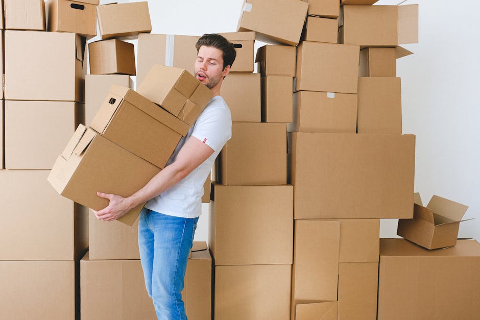 A man with dark hair, wearing a white t-shirt and blue jeans, is lifting a medium-sized cardboard box in an indoor setting surrounded by a large collection of stacked cardboard boxes of various sizes. Some boxes are sealed with packing tape, while others are open or partially open, revealing their corrugated texture. The man appears to be in the process of packing or preparing furniture for transport, which is part of a home relocation or moving process. Behind him, there are more cardboard boxes piled neatly against the wall, and the scene includes moving equipment such as a trolley or dolly to assist with heavy lifting. Natural lighting illuminates the space, indicating daylight coming through nearby windows. This visual showcases the packing and loading stages involved in house removals or furniture transport, aligning with services provided by Barnet Man and Van, a specialist in local removals across the Barnet area.
