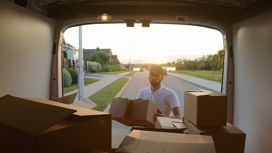 A man and a woman are inside a room surrounded by unpacked moving boxes made of cardboard, some labeled with handwritten notes such as 'books.' The woman is sitting on wooden flooring, wearing light-colored casual clothing and white socks, reading a book or manual. The man, dressed in a grey hoodie, is leaning on a box, attentively listening or observing. Several other packed boxes of various sizes are stacked around them, some on the floor and others against the window wall, with additional packing materials visible. The room features a large window with natural light illuminating the scene, dark curtains, and a neutral wall. This image depicts the home relocation process involving the unpacking and sorting of possessions, likely during a furniture transport or moving service. Barnet Man and Van's moving crews could be involved in this process as part of their house removals and packing services, preparing the property for further furniture transport or setup in the new residence.